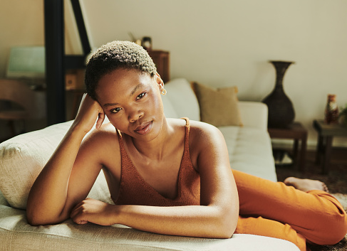Serious portrait of a young black woman on a sofa in her home sitting in a comfortable living room. Tired and exhausted young female from South Africa relaxing and sitting on couch in lounge of her house looking into the camera with copy space stock photo