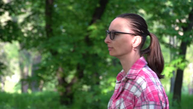 Woman in glasses and pink shirt walking in grove deciduous trees.