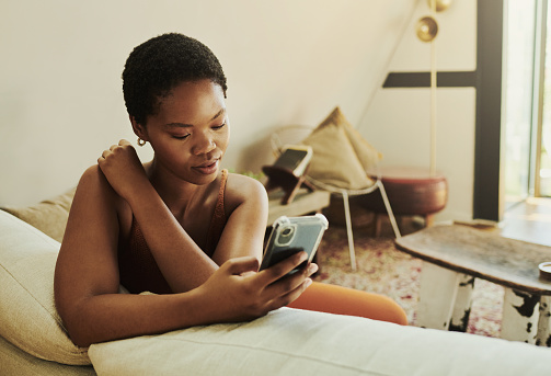 Young black woman comfortably sitting on her couch whilst reading a book on her mobile phone with copy space stock photo