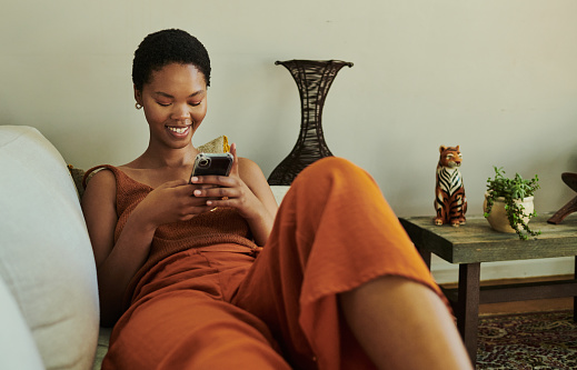 Young black woman laying on her back on her couch whilst scrolling through her mobile phone on social media smiling with copy space stock photo.