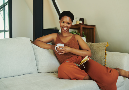 Young happy black woman smiling at the camera, sitting on a sofa or couch whilst casually dressed drinking coffee whilst having a book on her lap with copy space stock photo.