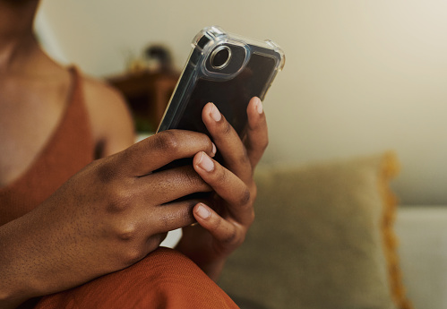 Unrecognized young black woman, sitting comfortably on her couch whilst texting on her mobile phone whilst casually dressed with copy space, stock photo.