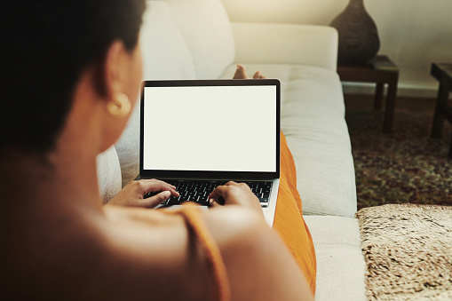 Young black unrecognized woman sitting with her back to the camera with her legs on her sofa or couch whilst typing on her laptop with copy space. stock photo