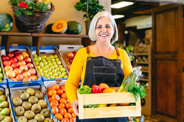 Female greengrocer working at the market holding a box containing fresh fruits and vegetables - Food retail concept stock photo
