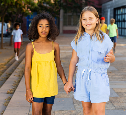Happy blonde preteen girl walking with her african american girl playmate along city street on summer day