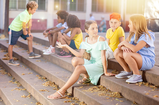 Group of multiracial boys and girls sitting on stairs outdoors and having conversation.