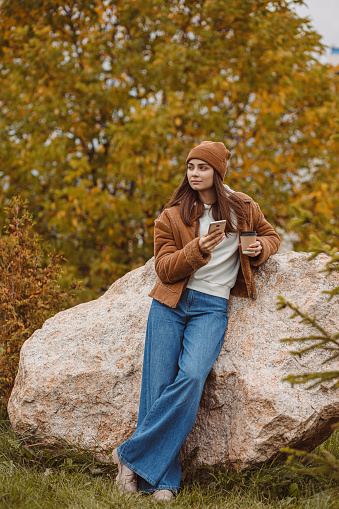 Thoughtful female hipster with paper cup of takeaway coffee standing in autumn park and using smart phone during weekend