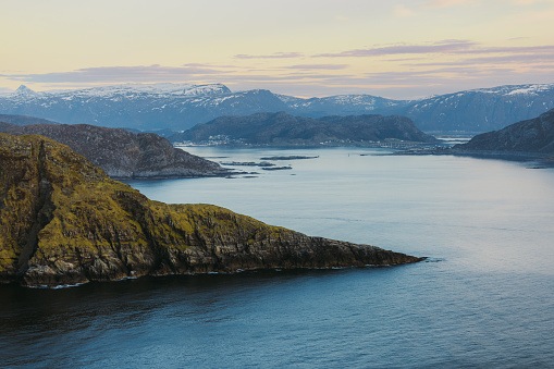 High-angle view of summer Scandinavian landscape with cliffs of the islands, snowcapped mountains and the ocean during sunset on Runde island, Western Norway