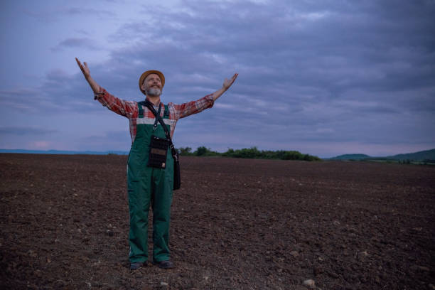 Cheerful farmer in green overalls, red checkered shirt, and straw hat in a plowed field before sunrise. He stands with his arms outstretched.