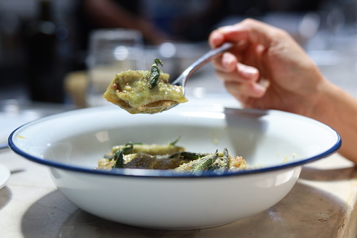 Cropped shot captures an Asian woman enjoying a bowl of spinach agnolotti filled with mortadella and ricotta, accompanied by a lemon zest butter sauce, in a restaurant.