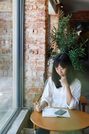 A woman sits in a cafe, checking travel information on her smartphone and simultaneously writing in a notebook to plan her trip.