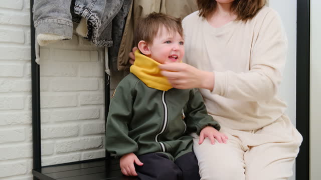 The parent puts a yellow scarf on their baby's neck before going out in the cold weather. A woman puts a yellow scarf on baby's head to keep them warm. Kid aged about two years (one year ten months)