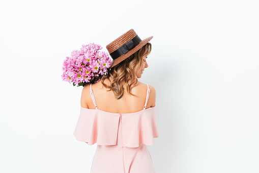 Romantic young woman wearing a pink dress and straw hat holding a bouquet of fresh flowers and smiling over her shoulder, rear view
