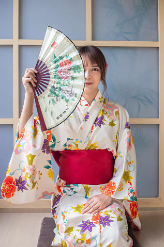 Young woman wearing Japanese traditional kimono holding paper fan sitting in Japanese style house