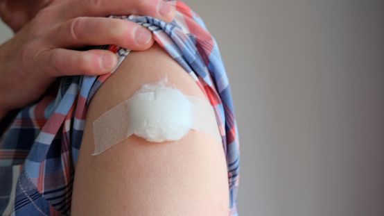 White male upper arm, shoulder or bicep, with small gauze or cotton pad plaster following vaccination injection (covid, flu) shirt sleeve lifted out of the way by hand. Copy space to right of arm.