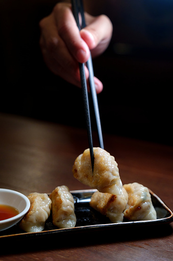 The close-up image captures a man skillfully using chopsticks to pick up a gyoza, delicately scooping the dumpling into the dipping sauce.