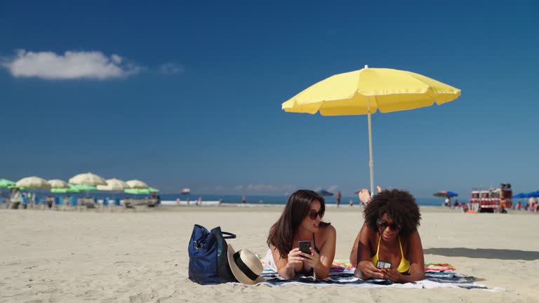 Two female friends chatting and using smarhphone while laying down at beach sunbathing