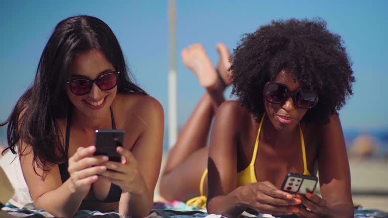 Two female friends chatting and using smarhphone while laying down at beach sunbathing