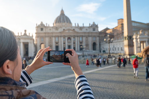 Vatican, December 23, 2022: Female tourist using smart phone for taking a photo of St. Peter's Cathedral, situated in the heart of Vatican City, a magnificent places visited by travelers from all over the world especially the ones visiting Rome and Italy