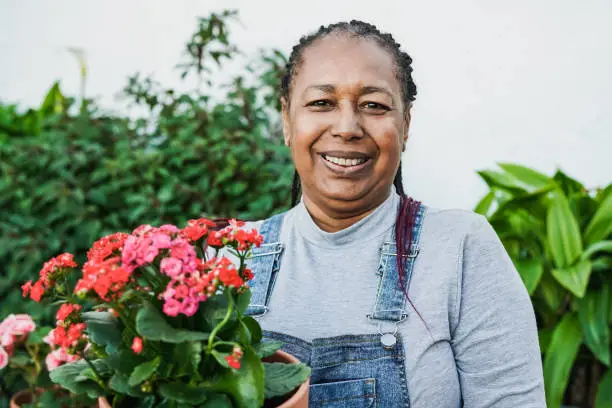 Photo of Happy african senior woman smiling on camera while holding flowers at home terrace - Gardening concept