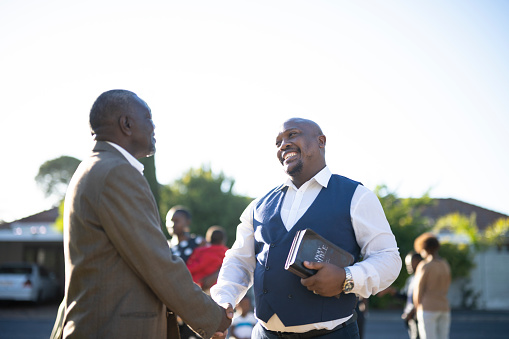 Two smiling senior men greet each other with a handshake as they arrive outside the  church, one man clutches his bible