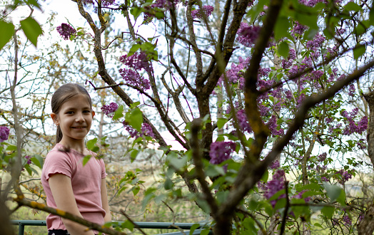 Beautiful happy smiling schoolgirl standing near blooming purple lilac flower tree.