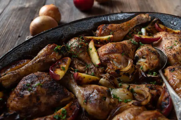 Delicious poultry dish with roasted chicken shanks and drumsticks. Cooked with apples and onions. Served ready to eat in rustic roasting pan on wooden table. Closeup
