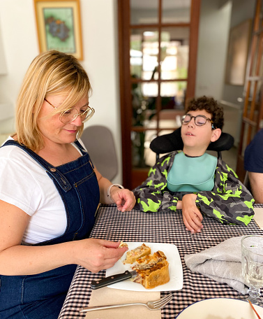 Mom feeding her son with cerebral palsy sitting in the wheelchair in the dinning room