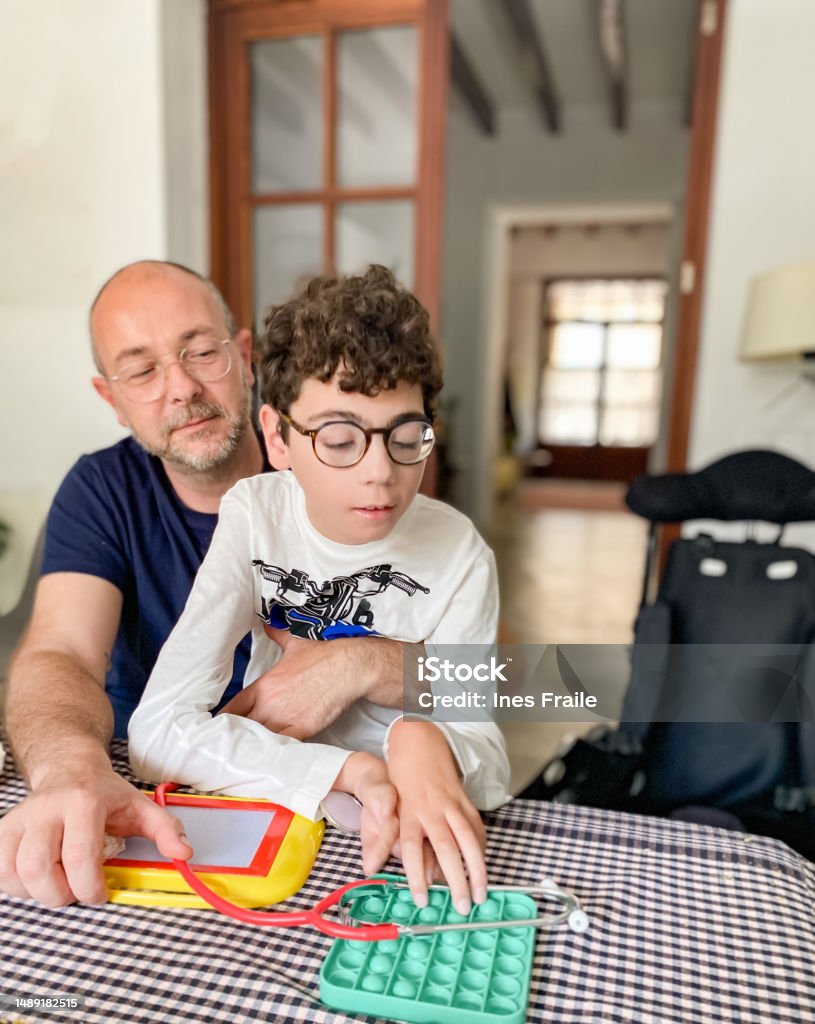 Father with his disabled son sitting on top playing with a push pop fidget toy Father with his disabled son sitting on top playing with a push pop fidget toy in a dinning room Autism Stock Photo