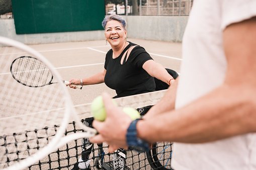 Disabled Latin woman practice wheelchair tennis with her coach outdoors. She has both artificial legs and disfigured left palm. Both dressed in casual sport clothes. Exterior of public tennis court in the public city park during the day.