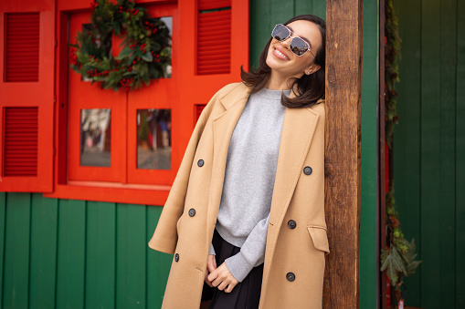 People, season and Christmas concept portrait of happy smiling middle age woman outdoors dressed stylish cream trench coat with Christmas festive decoration on background. Copy space