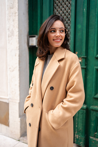 Friendly smiling attractive woman leaving building on background green hedge in the street of European city. Happy confident woman in glasses dressed stylish trench coat smiling looking at camera