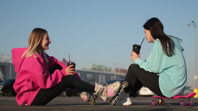 Skater girl friends drink coffee and talk sitting on asphalt