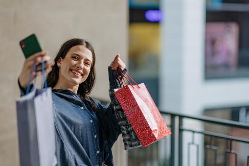 A young Caucasian woman is standing at the mall and cheerfully taking a selfie with the things she bought.