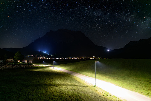 Nachtaufnahme: beleuchteter Fußweg nach Ehrwald vor der Zugspitze unter dem Sternenhimmel