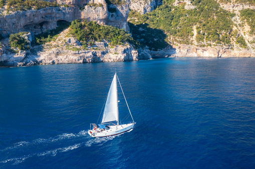 Aerial view of floating sailboat and rocky sea coast at summer sunny day. Top drone view of yacht, mountains, trees and blue water. Yachting in Sardinia, Italy. Tropical seascape with boat in ocean