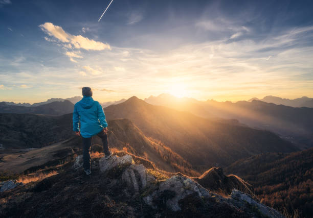 丘の上の石の上の男と、秋の色とりどりの夕日に霞の中の美しい山の谷。ドロミテ、イタリア。男、霧の中の山の尾根、オレンジ色の草と木々、秋の太陽と青い空。ハイキング - footpath single lane road sunrise landscape ストックフォトと画像