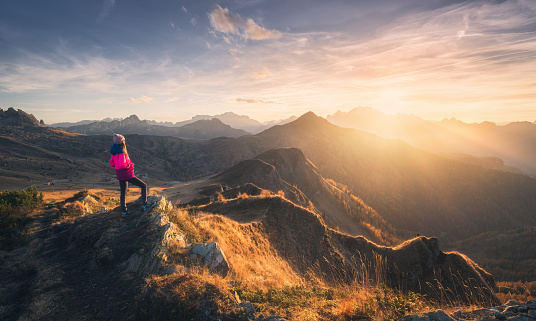 Young woman on mountain peak and beautiful mountain valley in haze at colorful sunset in autumn. Dolomites, Italy. Sporty girl, mountain ridges in fog, orange grass, trees, golden sun in fall. Hiking