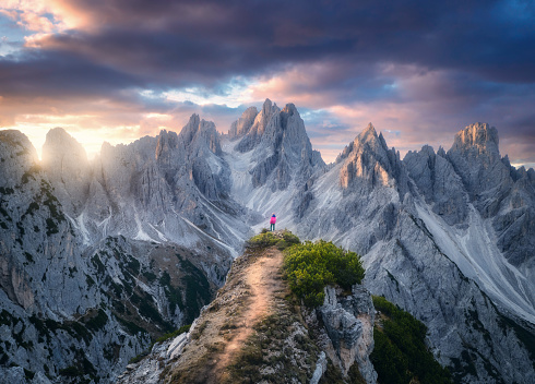 Aerial view of girl on the mountain peak and rocks at colorful sunset in summer. Tre Cime, Dolomites, Italy. Top drone view of woman on trail, cliffs, grass, cloudy sky, sunlight in spring. Hiking