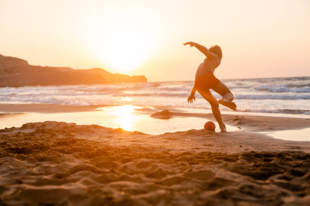 Cropped shot of the legs of youthful friends playing with ball together on the beach at sunset on a golden summer evening. Couple walking barefoot on sandy beach. They are walking, having fun.