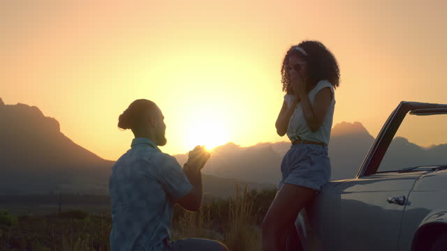 Man, woman and engagement proposal with sunset in summer by mountains, wow or road trip by car. Young woman, man and marriage offer in nature with convertible vehicle with ring, question and kneeling