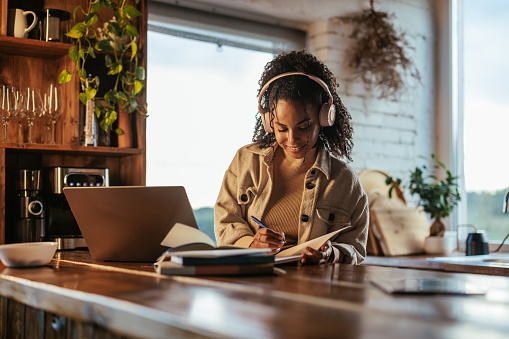 A young female student is at home studying at her laptop, catching notes in her notebook as she listen's to online classes.