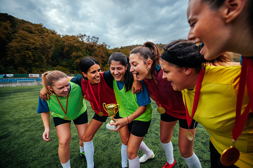 A group of young Caucasian female soccer club members are on the pitch celebrating after victory with a trophy.