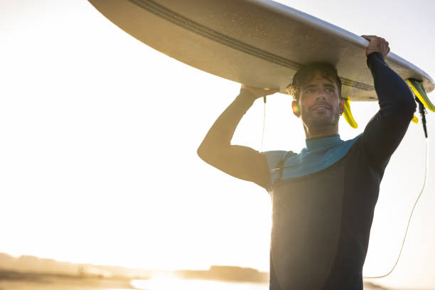 istock A surfer carries a surfboard walking along a sandy beach, with the sun setting in the background, casting a warm, golden light across the scene. 1489249724
