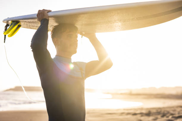 istock A surfer carries a surfboard walking along a sandy beach, with the sun setting in the background, casting a warm, golden light across the scene. 1489249728