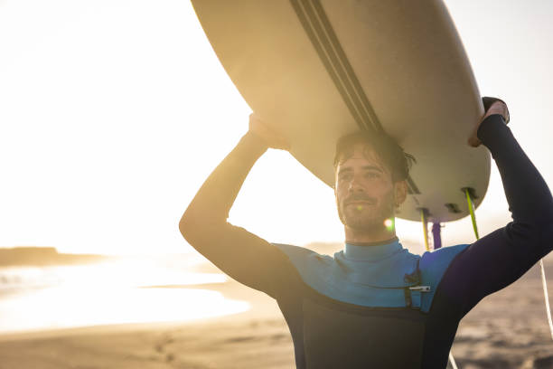 istock A surfer carries a surfboard walking along a sandy beach, with the sun setting in the background, casting a warm, golden light across the scene. 1489249745