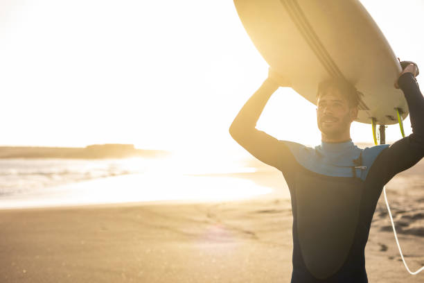 istock A surfer carries a surfboard walking along a sandy beach, with the sun setting in the background, casting a warm, golden light across the scene. 1489249750