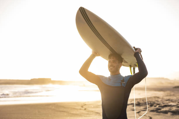 istock A surfer carries a surfboard walking along a sandy beach, with the sun setting in the background, casting a warm, golden light across the scene. 1489249755