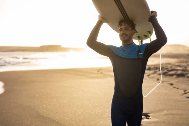 A surfer carries a surfboard walking along a sandy beach, with the sun setting in the background, casting a warm, golden light across the scene. stock photo