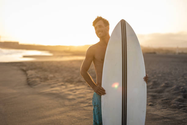 istock A surfer carries a surfboard walking along a sandy beach, with the sun setting in the background, casting a warm, golden light across the scene. 1489249835